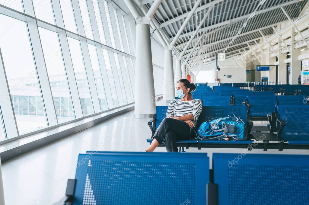 Lonely female solo traveler with backpack sitting in the empty airport passenger transfer hall in protective face mask and looking out large windows. Traveling in worldwide pandemic time concept image