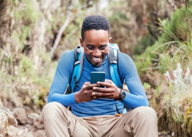 Genç Afrikalı etnik kökenli bir adamın cep telefonuyla sohbet etmesi ve ormanda yürüyüş yaparken neşeli bir şekilde gülmesi. Mutlu insanlar, ağ teknolojisi veya seyahat konsepti 