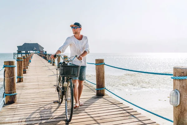 明るい夏の服を着た幸せな笑顔の裸足の男と木製の海の桟橋に自転車で歩くサングラスの肖像画 熱帯諸国での不注意な休暇のコンセプトイメージ — ストック写真
