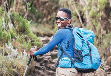 Neşeli gülümseyen, Afro-Amerikan etnik kökenli, güneş gözlüklü genç bir adamın portresi. Kilimanjaro 'daki Mweka kapısına giderken sırt çantasıyla yürüyüş yapıyor. Aktif tırmanış insanları konsepti.