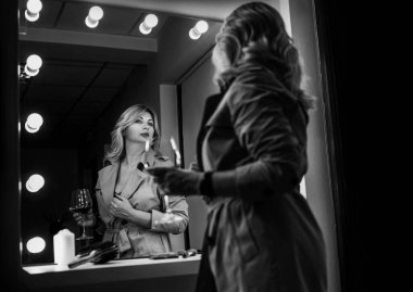 Orta yaşlı baştan çıkarıcı kadının portresi kırmızı şarap kadehiyle ayna yansımasına bakarak Trench ceketini takdir ediyor. İnsani duygu ya da giysi moda kavramı siyah beyaz görüntü.