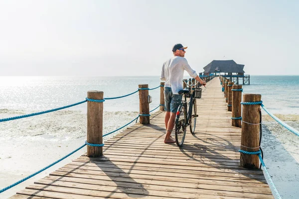 Back view portrait of a man dressed in light summer clothes, sunglasses with old bicycle on the wooden sea pier on the sandy Zanzibar beach.Careless vacation in the tropical countries concept image