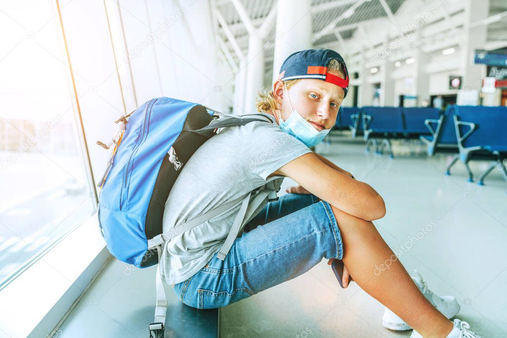 Portrait of lonely teen solo traveler with a backpack in empty airport passenger transfer hall in protective face mask and sadly looking at camera. Traveling in worldwide pandemic time concept image