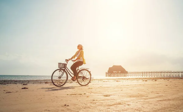 Young woman dressed light summer clothes riding old vintage bicycle with front basket on the lonely low tide ocean white sand coast on Kiwengwa beach on Zanzibar island, Tanzania.