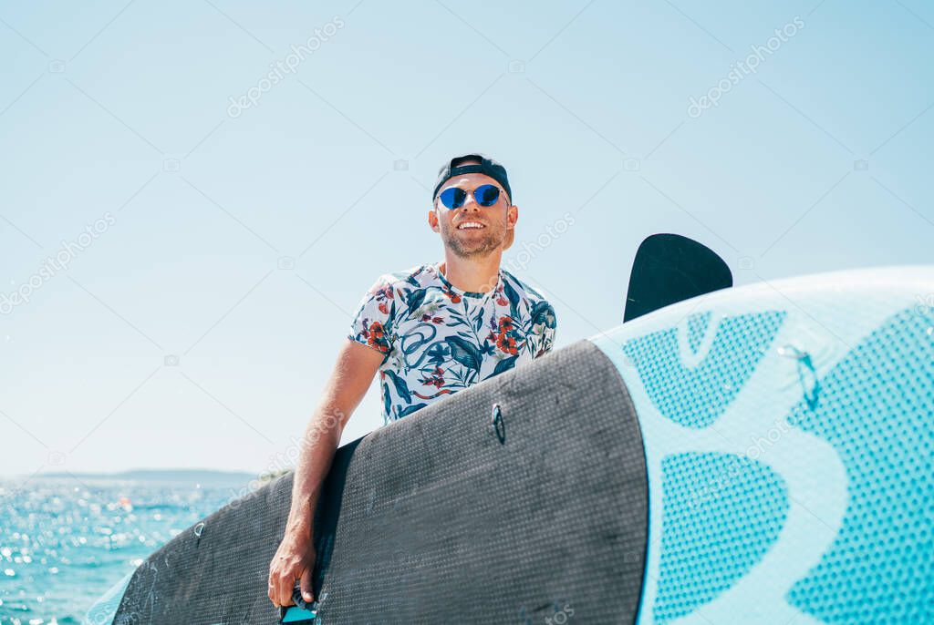 Smiling blonde surfer man in blue sunglasses and cap carrying the surfing board on the bright sunny day noon. Active people summer vacation time near the ocean concept image.
