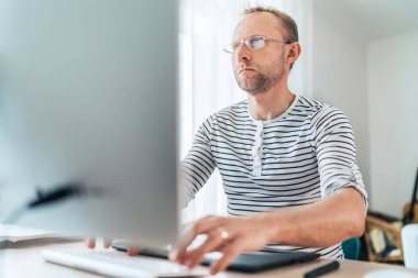 Gözlüklü orta yaşlı bir adam oturma odasında modern bir klavye bilgisayarı kullanarak mesaj yazıyor. Dünya çapında karantina zaman kavramı üzerine yazarlık, mesafe veya serbest çalışma 