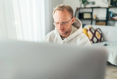 Gözlüklü orta yaşlı bir adam oturma odasında modern bir bilgisayar kullanıyor. Dünya çapında karantina zaman kavramı üzerine yazarlık, mesafe veya serbest çalışma 