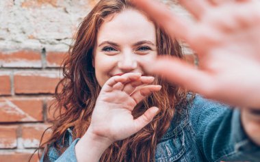 İçtenlikle gülümseyen kırmızı kıvırcık saçlı, beyaz saçlı, kırmızı ruj sürmüş, mavi dudaklı, kırmızı tuğladan bir arka planı olan ve fotoğraf çekmeyen genç bir kızın portresi..