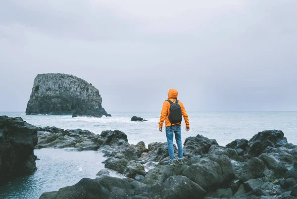 一位积极的背包客穿着橙色防水夹克 背包着背包 在岩石的马德拉岛海滨欣赏大西洋海湾美景 葡萄牙 — 图库照片
