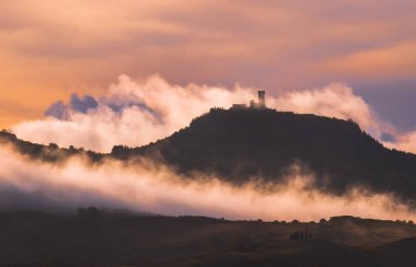 Rocca di Radicofani ana kulesi sabah sisiyle kaplı. Güneş ışığı, çimlerin ve tarlaların üzerinden akan kıvırcık bulutları kaplıyor. Tuscany tepelerinde ışık gölgeleri oluşturuyor..