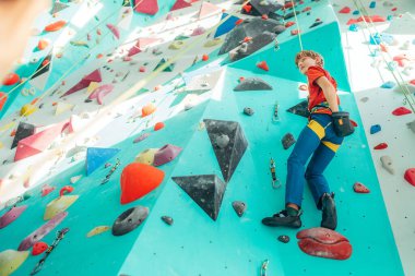 Kapalı alandaki genç çocuk duvardaki en üst halatla tırmanıyor. Çocuk tırmanma koşumu ve tebeşir tozu kullanarak tırmanırken biri onu yerden bağlamış. Etkin genç zaman harcama konsepti resmi.