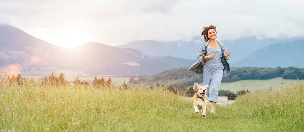 快乐地笑着跑着跑着的小猎犬的画像 伸出舌头 主人是女慢跑者 沿着草地小径跑着 与宠物在大自然中漫步 活跃的人的生活方式观念形象 — 图库照片