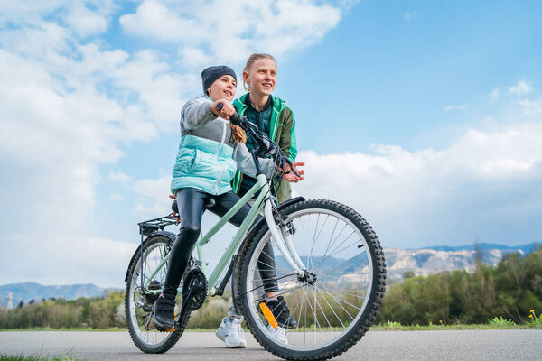 Brother helping to sister, supporting her and teaching doing first steps in riding. Kids on a bicycle path with wide blue cloudy sky background. Happy childhood concept image.