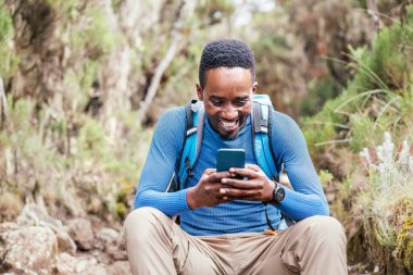 Genç Afrikalı etnik kökenli bir adamın cep telefonuyla sohbet etmesi ve ormanda yürüyüş yaparken neşeli bir şekilde gülmesi. Mutlu insanlar, ağ teknolojisi veya seyahat konsepti 