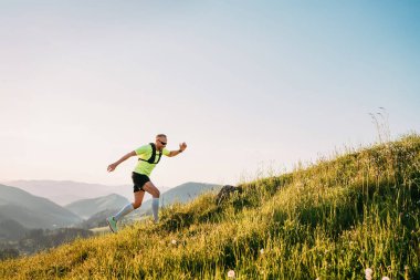 Orta yaşlı dağ koşucusu, güneş gözlüklü parlak bir sırt çantalı ve güneş gözlüklü bir tişört giymiş gün batımında tepelerden yukarı doğru koşuyor. Sportif aktif insanlar konsept imajı.