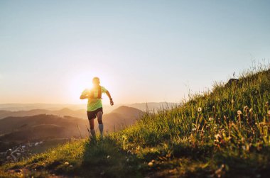 Aktif dağ koşucusu güneş gözlüklü sırt çantalı parlak bir tişört giyerek gün batımında pitoresk tepelerin yanından geçerek dayanıklılık maratonu yapıyor. Sportif aktif insanlar arka plan konsept görüntüsü.