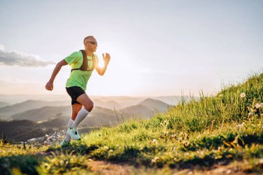 Orta yaşlı dağ koşucusu, güneş gözlüklü parlak bir sırt çantalı ve güneş gözlüklü bir tişört giymiş gün batımında tepelerden yukarı doğru koşuyor. Sportif aktif insanlar konsept imajı.