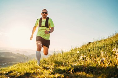 Aktif dağ koşucusu, güneş gözlüklü sırt çantalı parlak bir tişört giyerek gün batımında pitoresk tepelerde dayanıklılık maratonu yapıyor. Aktif sporcu imajı.