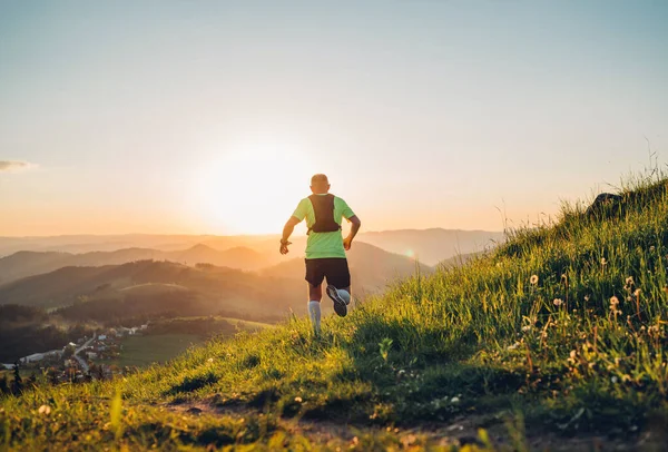 积极的山地小径跑步者穿着亮晶晶的T恤 背着背包跑耐力马拉松比赛 在日落时带着风景如画的小山向后看去 活泼活泼的人背光概念形象 — 图库照片