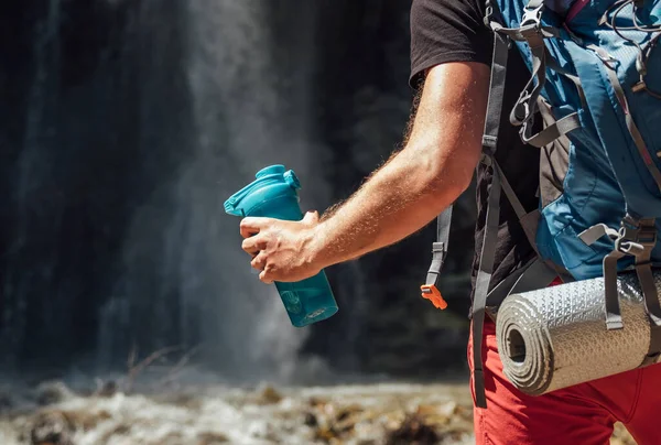 飲料水ボトルで手を閉じてください バックパックを着た男は山の川の滝の近くに滞在し 自然を楽しむ観光客のトレッキング服を着ています トレッキングのコンセプトイメージ — ストック写真