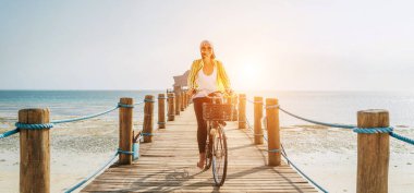 Hafif yaz kıyafetleri ve güneş gözlüğü takmış, tahta bir iskelede bisiklet süren ve kameraya bakan mutlu bir kadının portresi. Tropikal ülkelerde dikkatsiz tatil kavramı