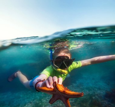 Genç bir çocuğun, elinde büyük turuncu bir denizyıldızı ile Zanzibar adasının yakınındaki mavi Hint Okyanusu dalgalarında dalış maskesiyle dalış yaparken sualtı görüntüsü. Egzotik tatil konsepti resmi.