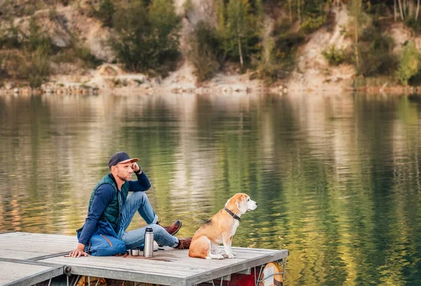 男犬の所有者と彼のビーグルの友人は 山の湖の木製の桟橋に座って 秋のシーズンの時間に歩いている間に風景を楽しんでいます 人間とペットのコンセプトイメージ — ストック写真