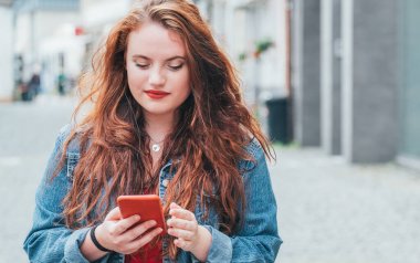 Teknoloji cihazları olan modern insanlar imaj kavramına sahipler. Kızıl bukleli uzun saçlı beyaz bir genç kızın portresi. Sokaklarda yürüyor ve modern bir akıllı telefon kullanarak internette geziniyor..