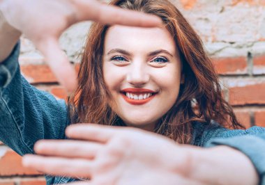 İçtenlikle gülümseyen kırmızı kıvırcık saçlı, beyaz saçlı, kırmızı rujlu, kırmızı dudaklı, kırmızı tuğladan bir arka planı olan ve 