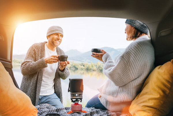 Car trunk view of chatting couple dressed warm knitted clothes enjoying gas stove prepared coffee and mountain lake view. Cozy early autumn couple auto traveling concept image.  