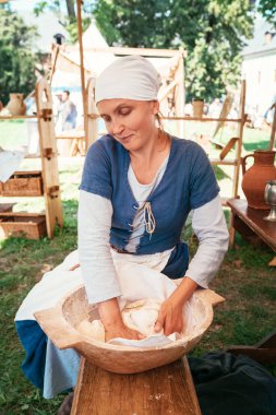 Geleneksel kostümlü bir ortaçağ kadını, ortaçağ festival çadırının altında kırsal ahşap masa, kil çömlek ve hasır sepetlerle çevrili tarihi canlandırma etkinliğinde ekmek hamuru hazırlıyor.