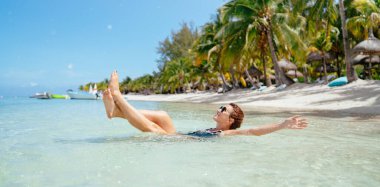 Güzel esmer kadın, siyah mayo giyip güneş gözlüğü takıyor sığ kumlu kumsal dalgalarında uzanarak egzotik lüks tatil yerleri ve şemsiyelerle dinleniyor. Egzotik tatiller ve seyahat konsepti