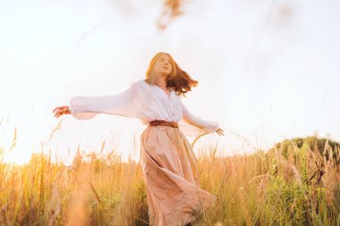 Beyaz bluz ve bej etekli genç bir kadın altın çayır boyunca özgürce yürüyor günbatımının keyfini çıkarıyor sıcak güneş ışığının barışçıl doğanın atmosferinin tadını çıkarıyor özgürlüğün farkındalığını ve doğayla uyum içinde olduğunu ifade ediyor.