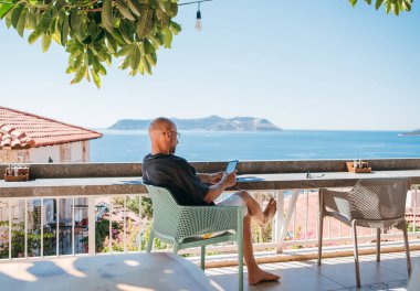 Kel adam güneşli bir otel terasında yaz tatilinin tadını çıkarıyor. Etrafı yeşilliklerle çevrili. Dijital okuyucu cihazıyla ilgili en son en çok satan e-kitabı okuyor, yaz tatilinin tadını çıkarıyor..