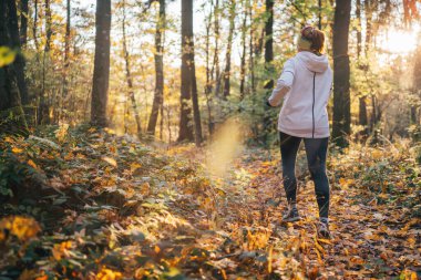 Sonbahar ormanlarında koşan genç bir kadın renkli yapraklarla çevrili aktif yaşam tarzının tadını çıkarıyor. Sabah sporu rutini. Doğa ile sağlıklı yaşam tarzı bağlantısı.