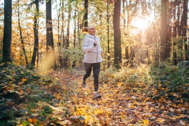 Ormanda koşu yapan orta yaşlı bir kadın renkli yapraklarla çevrili aktif yaşam tarzının tadını çıkarıyor. Sabah sporu rutin açık hava sağlığı, doğa konseptiyle sağlıklı yaşam tarzı bağlantısı