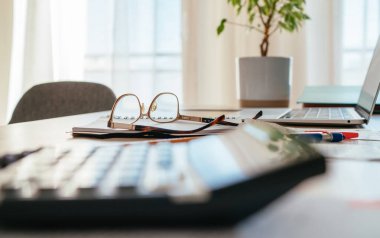 Gözlük ve hesap makinesi yakın plan ve modern dizüstü bilgisayarlı belgeler bir sonraki parlak gün ışığı penceresinde masa arkasında duruyor. İş, ev, para tasarrufu.