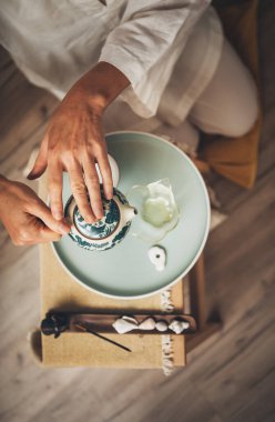 Erkek eller kil çaydanlıktan temiz cam sürahiye çay dökerek dingin çay ayini yaratıyor. Yumuşak ışık, ahşap çay tepsisi ve yeşillik dingin Çin seremonisi atmosferini canlandırıyor..