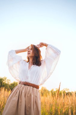 Beyaz bluz ve bej etekli genç bir kadın altın çayır boyunca özgürce yürüyor günbatımının keyfini çıkarıyor sıcak güneş ışığının barışçıl doğanın atmosferinin tadını çıkarıyor özgürlüğün farkındalığını ve doğayla uyum içinde olduğunu ifade ediyor.