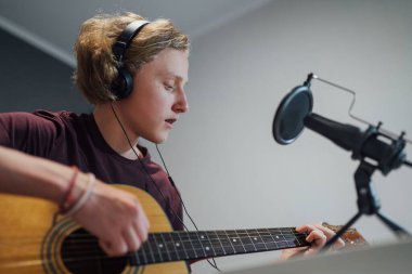 Kulaklıkla gitar çalan, mikrofon ve dizüstü bilgisayarla müzik kaydeden bir gencin ev ses stüdyosu portresi. Modern ses kayıt teknolojisi konsepti şık çocuk odasında.