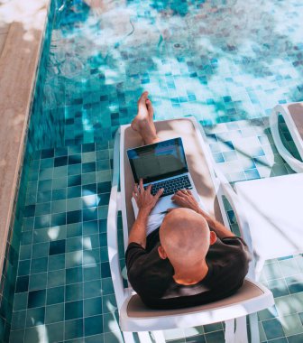 Serbest çalışan kel bir erkek CEO 'nun lüks otellerin havuzunda dizüstü bilgisayarla güneşlenerek çalışmasının en iyi görüntüsü. Uzak tropikal yaz tatilinde dijital göçebe şirketi başarıyla yönetiyor.