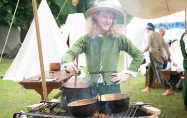 Ortaçağ hasır şapkası ve yeşil tunik aşçıları içinde gülümseyen genç erkek tarihi yeniden canlandırma etkinliğinde açık ateşte pişiyor. Yeniden yapılandırmalar, günlük yaşam ve geçmiş dönem geleneklerine dair değerli anlayışlar sağlar..