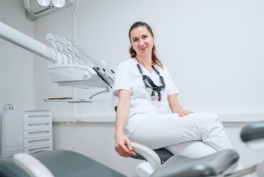 Modern diş sağlığı kliniğinde stomatoloji kürsüsünün yanında oturan beyaz önlüklü, gülümseyen genç dişçi kadın portresi. Sağlık ve sağlık sektörü konsepti.