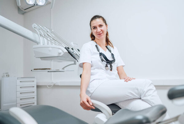 Portraait of sincerely smiling young dentist woman dressed white medical scrubs uniform sitting in modern dental clinic next to stomatology chair. Health care and medicare industry concept image.