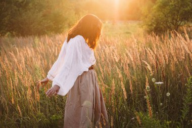 Beyaz bluzlu ve uzun etekli güzel bir kadın yaz mevsiminde huzurlu romantik yaşam tarzının tadını çıkarırken kırsal kesimdeki çayırlarda uzun ve kuru otların arasında yürüyor..