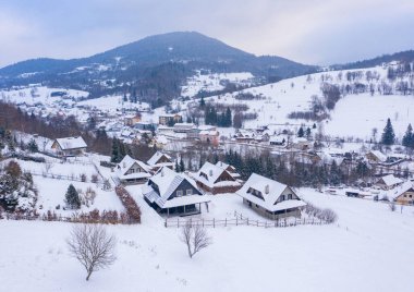 Slovakya 'daki Mala Fatra dağlarındaki karlı Terchova köyündeki geleneksel ahşap kulübelerin hava manzarasında beyaz kar kaplı tepeler ve bulutlu gökyüzü altında kırsal evler yer alıyor.