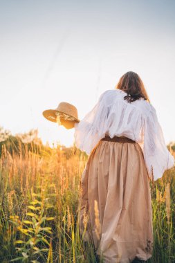 Özgür ruhlu kadın gün batımında altın çayırlarda yürüyor, elinde hasır şapkayla kırsal alanda huzurlu bir akşam gezintisi boyunca doğanın huzurunun tadını çıkarıyor.
