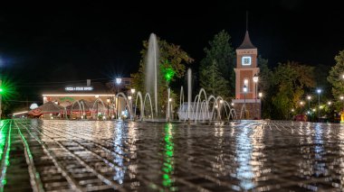 Bulgaristan 'ın Obzor kentindeki aydınlık çeşme ve saat kulesinin gece manzarası, ıslak kaldırımlar, atmosferik seyahat ve şehir merkezinde yakalanan turizm manzaraları yansımaları