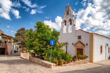 Margaritler, Yunanistan - 7 Eylül 2025: Çan kulesi, saat ve çiçek saksılarıyla bembeyaz yıkanmış kilise mavi gökyüzünün altında bulutlu Girit köyünde.