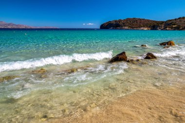 Yunanistan 'ın Girit kentindeki güzel Voulisma Sahili, turkuaz açık deniz, kumlu sahil ve kayalık sahili canlı mavi gökyüzü altında gösteriyor, tipik Akdeniz yaz manzarası.